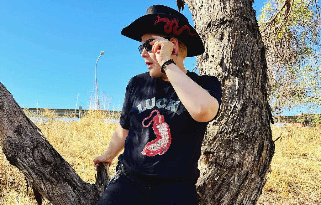 Person wearing a black t-shirt with a red graphic and text "Lucky", sitting against a tree in an outdoor setting.