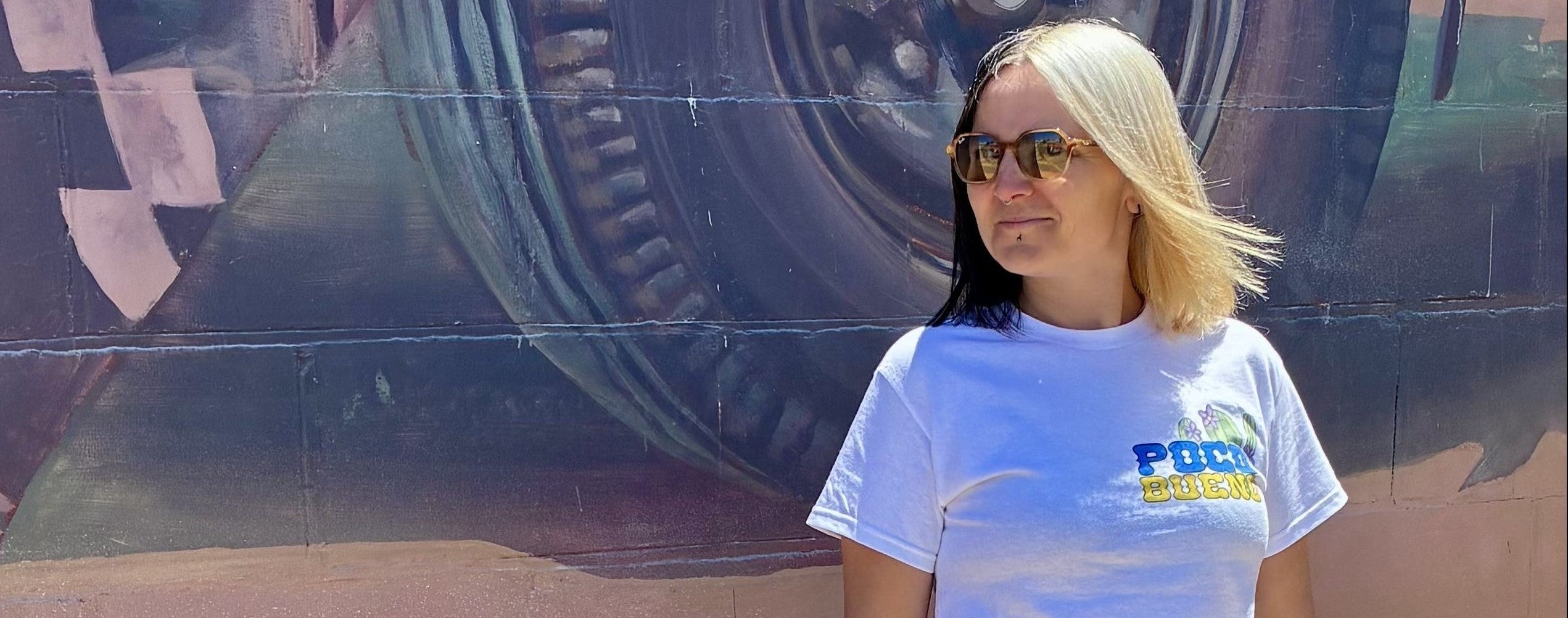 Woman wearing a white t-shirt with visible Poco Bueno branding in front of a large mural.